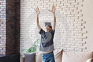 A teenage boy wearing glasses in a VR headset plays virtual games in his room. The young man raised his hands