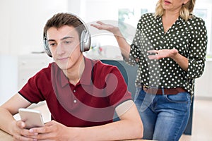 Teenage Boy Wearing Headphones And Using Mobile Phone Being Nagged By Mother At Home photo