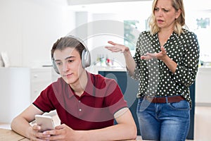 Teenage Boy Wearing Headphones And Using Mobile Phone Being Nagged By Mother At Home photo
