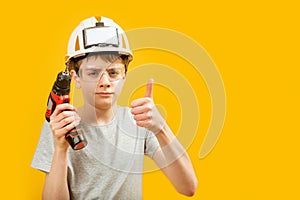 Teenage boy wears protective helmet and glasses holding drill and showing thumbs up, yellow background. Child as builder. Copy