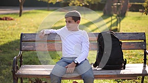 Teenage boy in white sweater sitting on the bench. Handsome school boy with backpack in autumn park