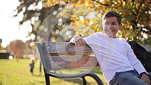 Teenage boy in white sweater sitting on the bench. Handsome school boy with backpack in autumn park