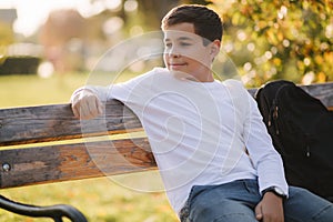 Teenage boy in white sweater sitting on the bench. Handsome school boy with backpack in autumn park