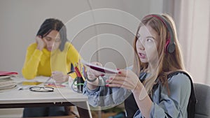 Teenage Caucasian girl in earphones throwing away notebook and singing. Cute teen resting as her African American friend