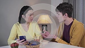 Teenage Caucasian son coming to smiling adult woman using phone and telling shocking news. Worried mother looking at boy