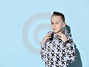 Teenage child with headphones on neck, in stylish hoodie and black bracelet. He smiling while posing against blue background