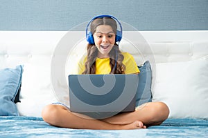 Teenage child in headphones relax on bed at home listen to music using laptop. Child in earphones browse internet on