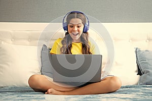 Teenage child in headphones relax on bed at home listen to music using laptop. Child in earphones browse internet on