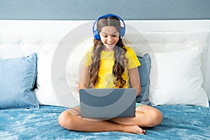 Teenage child in headphones relax on bed at home listen to music using laptop. Child in earphones browse internet on