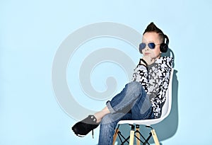Teenage child in headphones, sunglasses, blue jeans, hoodie, sneakers. Propping chin by fist, sitting on chair. Blue background