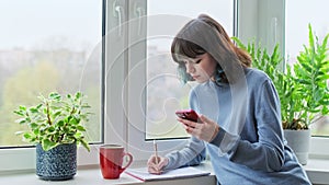 Teenage college student studying writing in notebook at home using smartphone