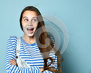 Teenage female in jeans overall, striped sweatshirt. She smiling, showing thumbs up, winking, posing on blue background. Close up