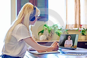 Teenage female student in headphones sitting at home using laptop talking to teacher