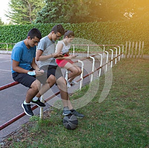 Teenage friends using a smartphone sitting on an outdoor playground fence - Young Mobile Smartphone Addicts