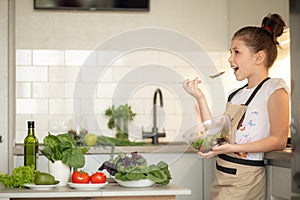 A teenage girl in apron prepared a salad on her own in the home kitchen. She holds a bowl of cooked salad and tastes it