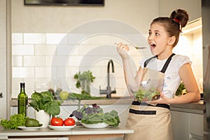 A teenage girl in apron prepared a salad on her own in the home kitchen. She holds a bowl of cooked salad and tastes it