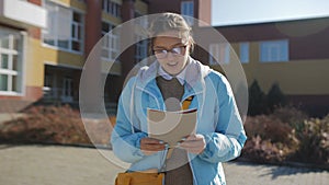 A teenage girl in a blue jacket and glasses on the background of an educational institution with a summary in her hands.