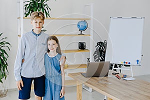 Teenage girl and boy standing near table with laptop in classroom looking to the camera. Teen schoolboy and schoolgirl do project