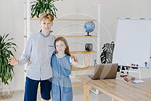 Teenage girl and boy standing near table with laptop in classroom looking to the camera. Teen schoolboy and schoolgirl do project