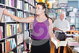 Teenage girl is choosing textbook for preparing to exam in bookstore.