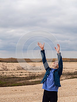 A teenage girl in a denim jacket looks up and stretches her arms to the sky, standing on the sandy seashore against the background