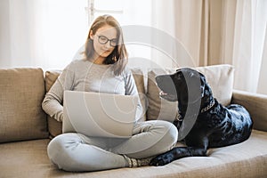 Teenage girl with a dog sitting on a sofa indoors, working on a laptop.