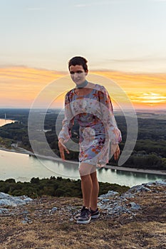 teenage girl in dress, with short-cropped hair, on top cliff, on river bank, at sunset