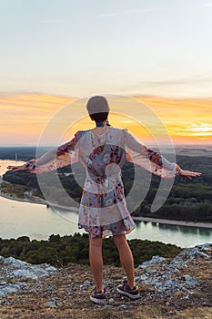 teenage girl in dress, with short-cropped hair, on top cliff, on river bank, at sunset