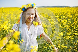 Teenage girl in dress and Ukrainian wreath with lentims on head, in rapeseed field under a clear blue sky