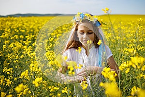 Teenage girl in dress and Ukrainian wreath with lentims on head, in rapeseed field under a clear blue sky