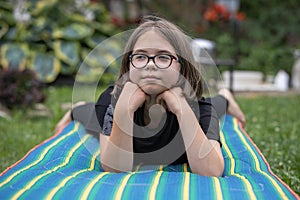 A teenage girl with glasses lies on a mattress on a lawn in a summer garden and thinks about something