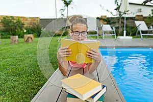 Teenage girl in glasses reads a book, background swimming pool, lawn near the house. School, education, knowledge, adolescents.