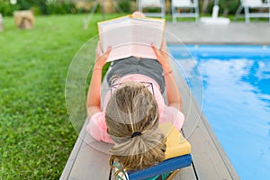 Teenage girl in glasses reads a book, background swimming pool, lawn near the house. School, education, knowledge, adolescents