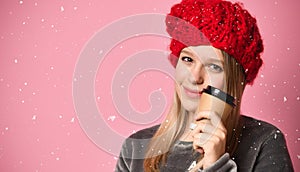 Teenage girl in gray pajamas, red hat. She is holding a paper cup and going to drink, posing on pink background. Close up