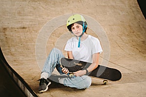 Teenage girl in helmet and stylish clothes posing on half pipe ramp an outdoor skate park. Beautiful kid female model skateboarder