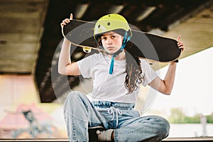 Teenage girl in helmet and stylish clothes posing on half pipe ramp an outdoor skate park. Beautiful kid female model skateboarder