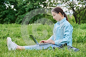Teenage girl high school student using laptop while sitting on green grass