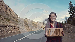 Teenage Girl Holding An Adventure Sign On A Mountain Road, Smiles And Laughs. Hitchhiking in search of adventure.