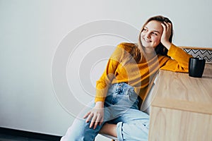 Teenage girl in home interior looks left side, lean on wooden table with black cup of coffee and sitting on white modern chair. Be