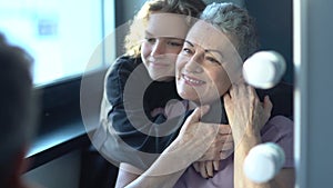 A teenage girl hugs her beautiful gray-haired grandmother sitting by the mirror. Two generations of one family, age