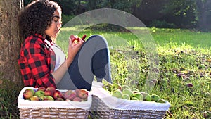 Teenage girl leaning against a tree in an orchard eating an apple using her mobile cell smart phone