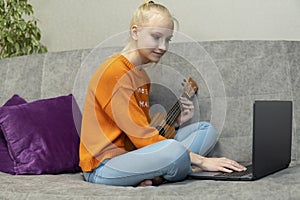 Teenage girl learns to play the ukulele and looks into her laptop.