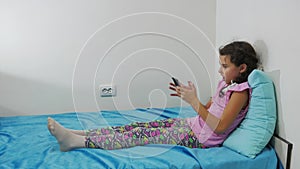 Teenage girl lies on a bed with a smartphone playing in an online game. Little girl surfing the Internet