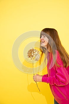 Teenage girl with long dark hair in pink sweater looks away and laughs, holding flower in hands. Bright photo, happiness, joy