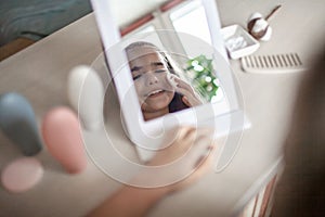 Teenage girl with long hair applying facial cosmetic mask before mirror, beauty salon at home