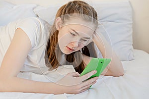 A teenage girl with long hair in a white T-shirt lies with a tablet in her hands on a white bed. The concept of adolescents`