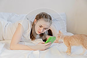 A teenage girl with long hair in a white T-shirt lies with a tablet in her hands on a white bed. The concept of adolescents`