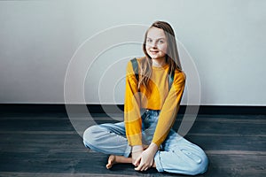 Teenage girl looking at camera and smiles sitting on wooden floor. Beautiful student girl in yellow sweater and blue jeans hold bo