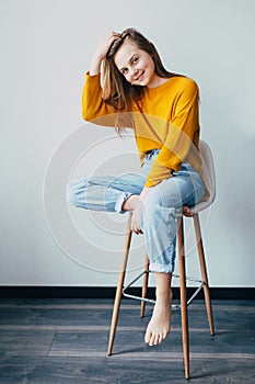 Teenage girl looks at camera with hand in her hair, sitting on white modern chair with one leg raise up. Beautiful girl in yellow