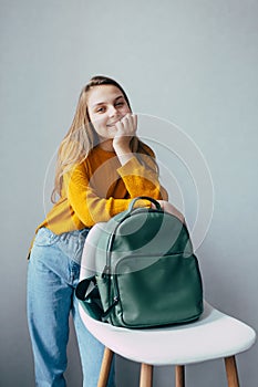 Teenage girl looks at camera .lean on hand and white modern chair with leather rucksacks. Beautiful girl in yellow sweater and blu